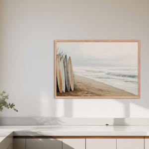 May include: A framed print featuring a beach scene with surfboards. The image shows several surfboards leaning against each other on a sandy beach. The ocean waves are visible in the background, with a light, neutral color palette.