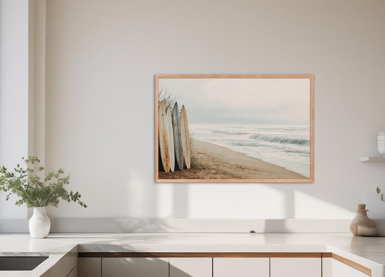 May include: A framed print featuring a beach scene with surfboards. The image shows several surfboards leaning against each other on a sandy beach. The ocean waves are visible in the background, with a light, neutral color palette.