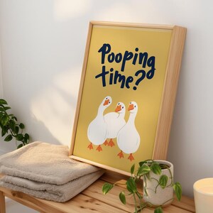 May include: A framed print with a yellow background and the text "Pooping time?" in navy blue. The print features three white geese with orange feet. The frame is made of light-colored wood. The print is displayed on a wooden bench with a folded towel and a small potted plant.