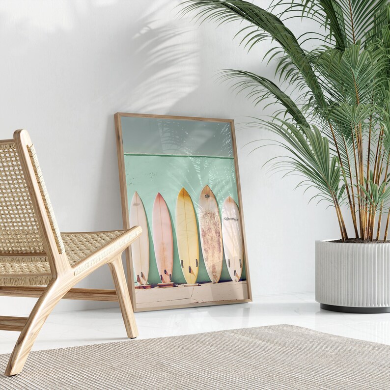 May include: A framed coastal-themed print featuring five vintage surfboards lined up against a teal wall. The surfboards are various pastel colors, including yellow, orange, and pink. The wooden frame complements the beachy aesthetic. The print is displayed in a room with a light-colored woven chair and a potted palm plant, creating a serene and stylish atmosphere.
