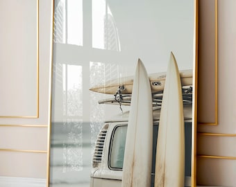 May include: A vintage white van with two long, white surfboards strapped to the roof. The van is parked in front of a blurry background of a beach and ocean.