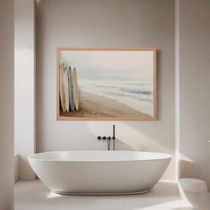 May include: A framed print of a beach scene with surfboards leaning against each other. The artwork is displayed above a white oval bathtub in a modern bathroom setting. The color palette is neutral with soft tones.