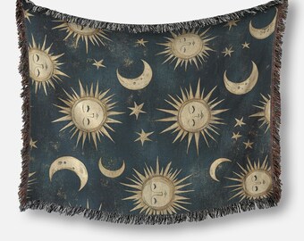 May include: A dark blue throw blanket featuring a celestial pattern of smiling suns, crescent moons, and stars in a vintage style. The blanket has a fringed edge and is made of a soft, woven material. The suns and moons are in a cream color.