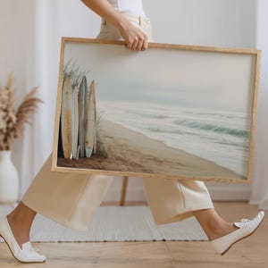 May include: A framed print featuring a serene beach scene with surfboards. The artwork depicts a sandy beach, ocean waves, and a cloudy sky. The surfboards are leaning against the dunes, adding a coastal theme to the artwork.