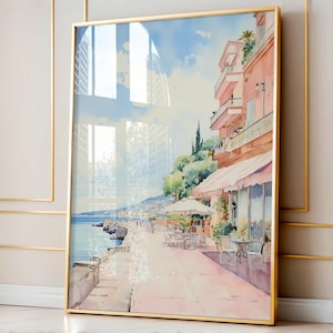 May include: A watercolor painting of a pink building with a cafe on the ground floor. The cafe has a pink awning and tables and chairs set up outside. The building is on a street that leads to a beach. The painting is framed in a gold frame.