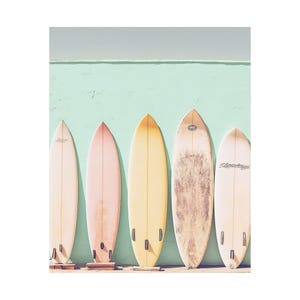 May include: Five pastel-colored surfboards stand against a light teal wall.  From left to right, the surfboards are off-white, light pink, pale yellow, beige with darker markings, and light peach.  The boards are similar in shape and size, with subtle variations in color and markings.  'Channel Islands' is visible on one surfboard.