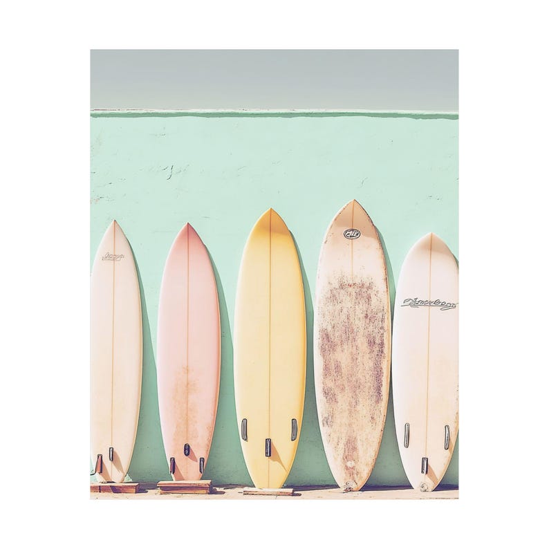 May include: Five pastel-colored surfboards stand against a light teal wall.  From left to right, the surfboards are off-white, light pink, pale yellow, beige with darker markings, and light peach.  The boards are similar in shape and size, with subtle variations in color and markings.  'Channel Islands' is visible on one surfboard.
