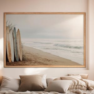 Puede incluir: Una impresión enmarcada que presenta una escena de playa con tablas de surf. Cuatro tablas de surf desgastadas se apoyan contra hierba alta en una playa de arena. Las olas del océano rompen bajo un cielo brumoso, creando una estética costera serena.