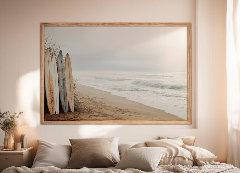 May include: A framed print featuring a beach scene with surfboards. Four weathered surfboards lean against tall grass on a sandy beach. The ocean waves roll in under a hazy sky, creating a serene coastal aesthetic.