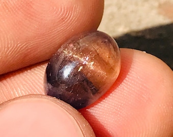 May include: A polished, oval-shaped gemstone with a mix of dark purple and golden-brown hues. The stone has a smooth, reflective surface, and is held between two fingers. The gem appears to be a sunstone.