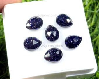 May include: Six dark blue teardrop-shaped gemstones with sparkling inclusions are displayed on a white surface. The gemstones are faceted, catching the light. The text "zsgemstones" is visible on the white surface. The gemstones are set against a green background.
