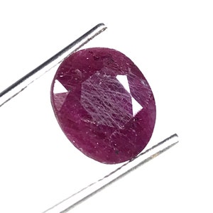 May include: A deep red oval-shaped gemstone with a faceted cut. The gemstone is held in a pair of tweezers.