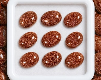 May include: A white square display tray containing nine oval-shaped, polished goldstone cabochons. Each cabochon features a deep reddish-brown base color with numerous embedded metallic copper-colored flecks that create a sparkling, glittery appearance.