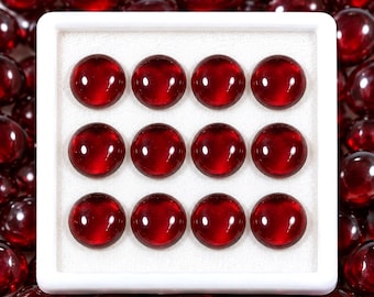 May include: A square white display box containing twelve round, deep red cabochon gemstones arranged in a three-by-four grid. The stones have a smooth, polished surface with a glossy finish, reflecting light clearly against the white foam insert.