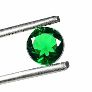 May include: A round, faceted, vibrant green gemstone held by silver-toned metal tweezers. The stone features a brilliant cut with multiple triangular and kite-shaped facets that reflect light, creating a clear, transparent, and deep emerald green appearance.