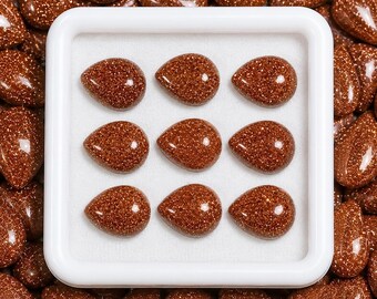 May include: A collection of nine teardrop-shaped goldstone cabochons arranged in a three-by-three grid within a white square display case. The stones are a deep reddish-brown color with a sparkling, metallic glitter effect throughout the surface.
