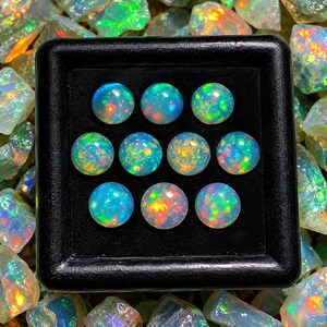 May include: A collection of nine round, cabochon-cut opals arranged in a three-by-three grid inside a black display box. Each opal exhibits a translucent, milky base with vibrant flashes of green, orange, yellow, and blue play-of-color.