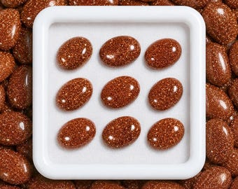 May include: Nine oval-shaped goldstone cabochons with a deep reddish-brown hue and sparkling metallic inclusions are arranged in a three-by-three grid inside a square white display tray, surrounded by a loose collection of identical stones.