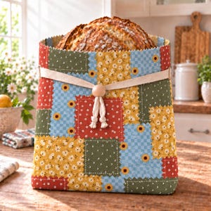 May include: A fabric bread bag featuring a patchwork design of floral and checkered patterns in red, yellow, green, and blue. The bag is cinched at the top with a beige fabric tie and a single wooden bead, holding a loaf of crusty bread.
