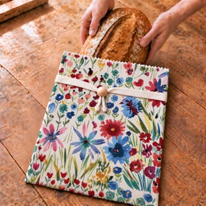 May include: A rectangular fabric bread bag featuring a colorful watercolor floral print with red, blue, pink, and green flowers on an off-white background. The bag is secured with a cream-colored fabric tie and a single round wooden bead.