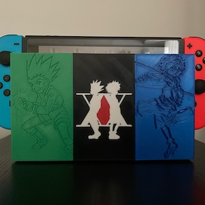 Peut inclure: Une coque Nintendo Switch en 3D imprimée en vert, noir et bleu avec un design Hunter x Hunter. La coque présente les personnages Gon Freecs, Killua Zoldyck et le logo Hunter x Hunter.