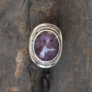 Może przedstawiać: Srebrny pierścionek z fioletowym owalnym kamieniem szlachetnym osadzonym w plecionym srebrnym obramowaniu.