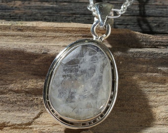 Puede incluir: Un colgante de plata con un cristal transparente de forma ovalada. El cristal tiene un aspecto nublado y lechoso con líneas internas visibles.