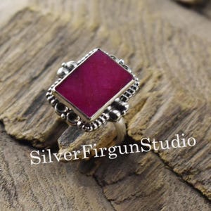 May include: A silver ring featuring a large, square, magenta gemstone. The ring has an ornate, textured setting with decorative details surrounding the stone. The text "Silver Firgun Studio" is visible on the wooden surface.