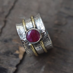 May include: A silver and gold spinning ring with a red gemstone. The ring has a textured design and is set with a round, faceted ruby.