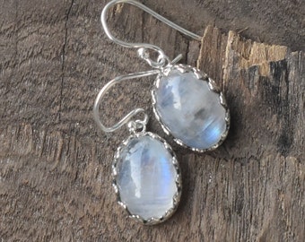 May include: A pair of silver earrings with oval-shaped moonstone gemstones. The stones have a milky white color with a hint of blue.