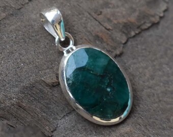 May include: A silver pendant featuring a large, oval-shaped emerald gemstone. The emerald is a deep green colour and has a faceted cut. The pendant is on a wooden surface.