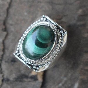Könnte beinhalten: Ein silberner Ring mit einem grünen Malachit-Stein, der in einer detaillierten ovalen Fassung sitzt. Der Ring hat eine strukturierte Band mit kleinen Perlen.