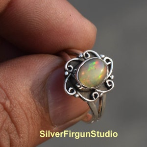 Puede incluir: Un anillo de plata con una piedra de ópalo ovalada engastada en un delicado diseño floral. El ópalo tiene una base blanca lechosa con destellos de verde, rojo y naranja.