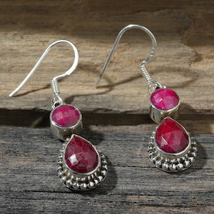 Może przedstawiać: Para srebrnych kolczyków z dwoma czerwonymi kamieniami szlachetnymi w kształcie kropli. Kolczyki mają kształt łzy i są ozdobione małymi srebrnymi koralikami.