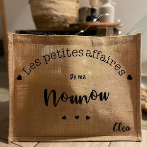 Peut inclure: Un sac fourre-tout rectangulaire en toile de jute beige avec le texte noir Les petites affaires De ma Nounou, des icônes de petits cœurs et le prénom Clea imprimé en bas à droite. Le sac présente une texture tissée et une forme structurée.