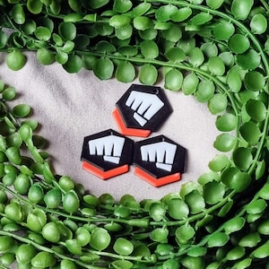 K&ouml;nnte beinhalten: Drei schwarze und wei&szlig;e sechseckige Anh&auml;nger mit einem Faust-Symbol in der Mitte. Die Anh&auml;nger haben einen roten Rand.