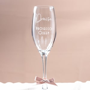 May include: A clear glass champagne flute with the text "Denise Prosecco Queen" etched on the side. The flute is tied with a pink ribbon.