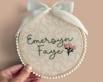 May include: A circular embroidery hoop containing fabric with a light pink vine pattern. The name Emersyn Faye is stitched in dark green thread, accompanied by a small cluster of three pink embroidered flowers and a white ribbon bow.