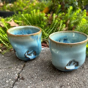 May include: Two handmade ceramic cups with a light blue glaze and brown accents. The cups have an organic shape and a textured surface. Each cup features a decorative brown design on the side. The cups are displayed outdoors.