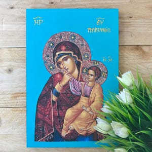 Può includere: Icona religiosa raffigurante la Vergine Maria con il bambino, su sfondo blu brillante. L'icona presenta scritte dorate e dettagli intricati. Le figure sono vestite con abiti tradizionali.