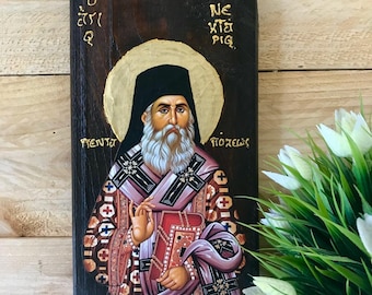 May include: Hand-painted icon of Saint Neophytos on a wooden panel. The icon depicts the saint with a white beard and a pink robe, holding a book. The background is a dark brown wood with gold accents.