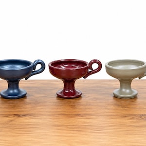 May include: Three ceramic chalices in a row, each with a handle. The chalices are in shades of blue, burgundy, and beige. They are placed on a wooden surface against a white background.