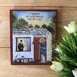 Könnte beinhalten: Religiöse Ikone, die zwei Figuren vor einem weißen Gebäude mit roter Tür zeigt. Die Ikone hat griechischen Text oben und ist von einem dunkelroten Rahmen umgeben. Der Hintergrund zeigt Bäume und einen Himmel.