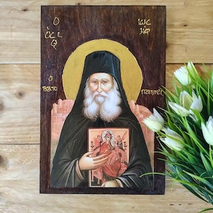 Pode incluir: Um ícone retangular de madeira apresentando um monge barbudo com hábito preto, segurando um ícone menor da Virgem com o Menino. O fundo é de madeira escura com um halo dourado e letras gregas. A obra de arte exibe uma cena religiosa detalhada.