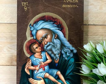 May include: A religious icon on a wooden panel. The image depicts an older man with a long, blue beard holding a child. The child is wearing blue clothing and holding a scroll. The background is a warm brown with gold accents and text.