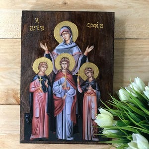 May include: Wooden icon featuring a religious image. The artwork depicts a central figure with arms raised, surrounded by three figures, all with halos. The figures are dressed in robes of various colors, including pink, blue, and black. Gold lettering is visible at the top.