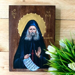 Pode incluir: Ícone religioso em painel de madeira. A imagem retrata um homem com longa barba branca e túnica azul escura, segurando um pergaminho. Uma auréola dourada envolve sua cabeça. O fundo é marrom escuro.