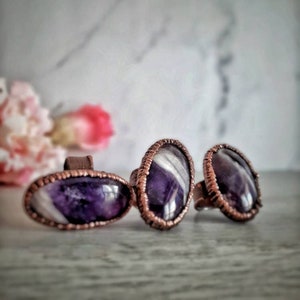 May include: Three rings with oval-shaped amethyst stones set in copper wire. The amethyst stones have a purple and white striped pattern.