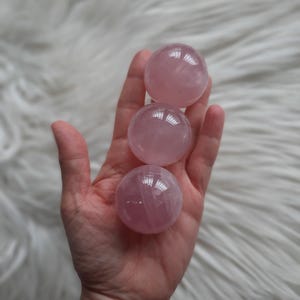 Può includere: Tre sfere di quarzo rosa, ciascuna con una superficie liscia e lucida, sono tenute in una mano. Le sfere sono di dimensioni diverse, con la sfera più grande in alto e la sfera più piccola in basso.