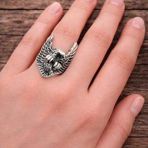Può includere: Anello color argento con design ad aquila. L'anello presenta una testa d'aquila dettagliata con ali spiegate. Il gioiello è indossato su un dito, mettendo in risalto l'intricata lavorazione e l'estetica audace del pezzo.